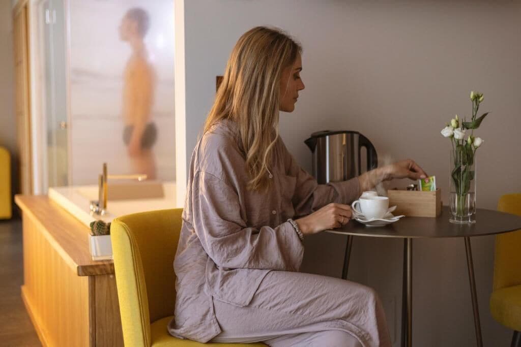 A person seated at a table with a cup and a vase of flowers, in a room with a wooden counter and a view of a person outside.