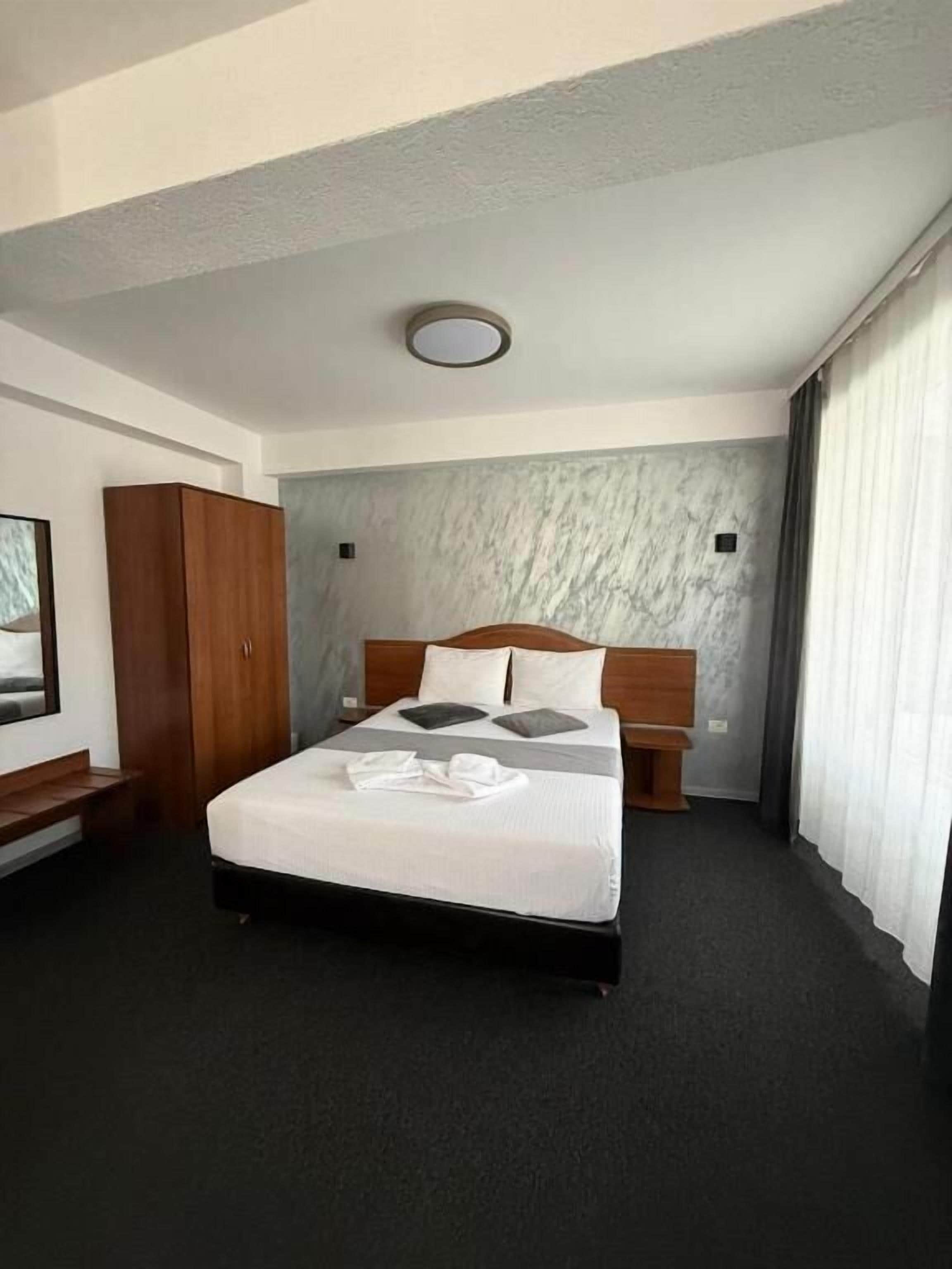 A hotel room with a bed, a wooden wardrobe, a nightstand, and a window with curtains.