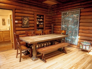 Een houten eetkamer met een grote tafel, bankjes en stoelen. Er staat een boekenkast met diverse spullen en er hangt een wandtapijt aan de muur.