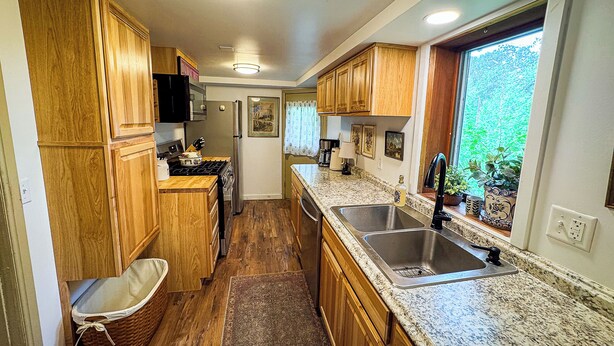 A compact kitchen with wooden cabinets, a stainless steel sink, and a window providing a view of greenery outside. - Storybook stone & log cabin with barrel sauna, 2 min to the beach (Herbster)