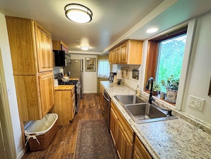 A compact kitchen with wooden cabinets, a stainless steel sink, and a window providing a view of greenery outside. - Camp Clover | Storybook Cabin Near Herbster Beach (Herbster)