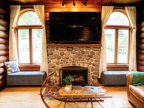A log cabin interior with a stone fireplace, a television mounted above it, and seating areas with windows. - Camp Clover | Storybook Cabin Near Herbster Beach (Herbster)