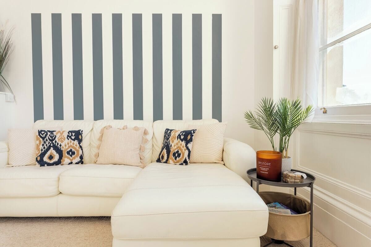 Un salon avec un canapé blanc, des coussins à motifs, une table d’appoint avec une plante et un mur à rayures verticales.