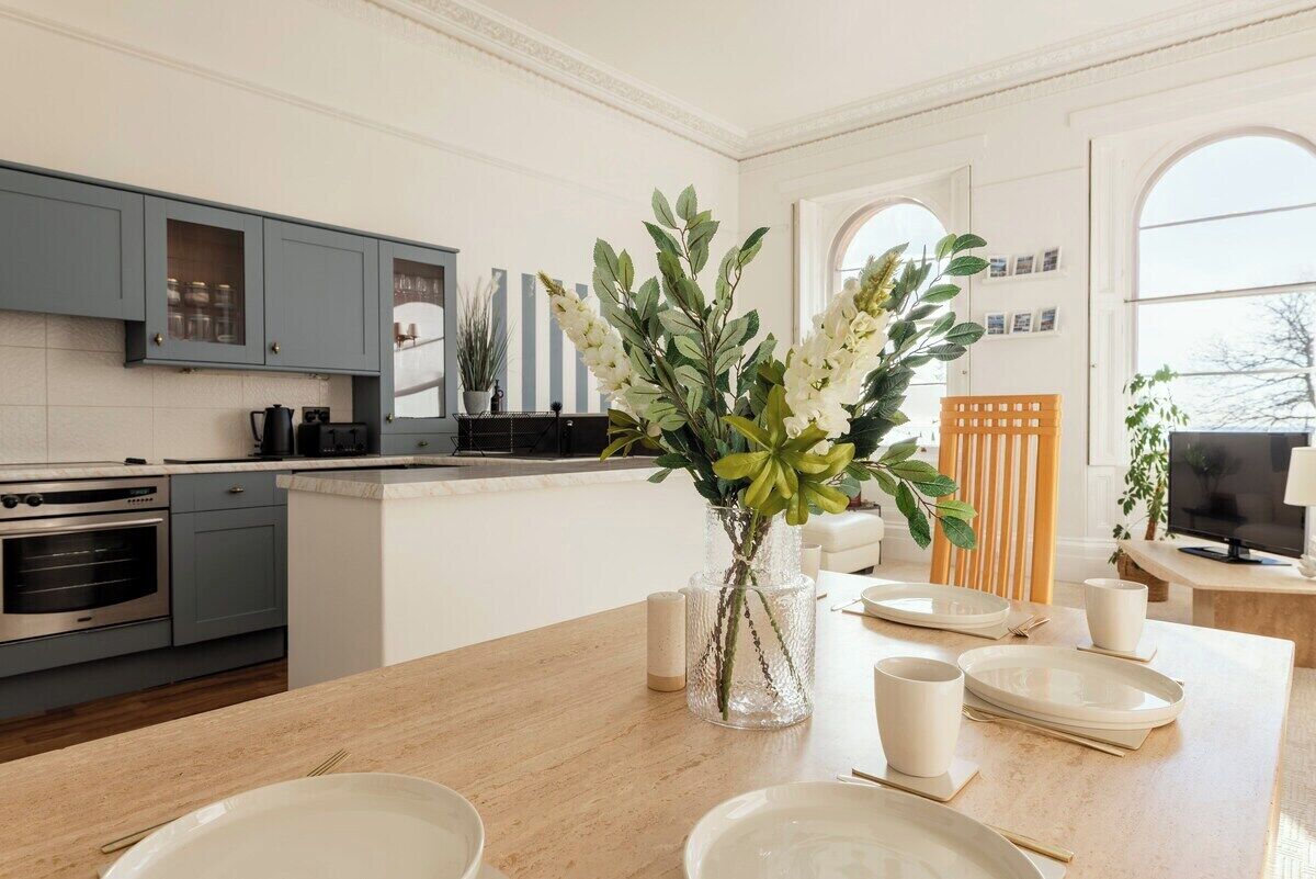 A modern kitchen with grey cabinets, a wooden dining table set with plates and cups, and a vase with flowers.