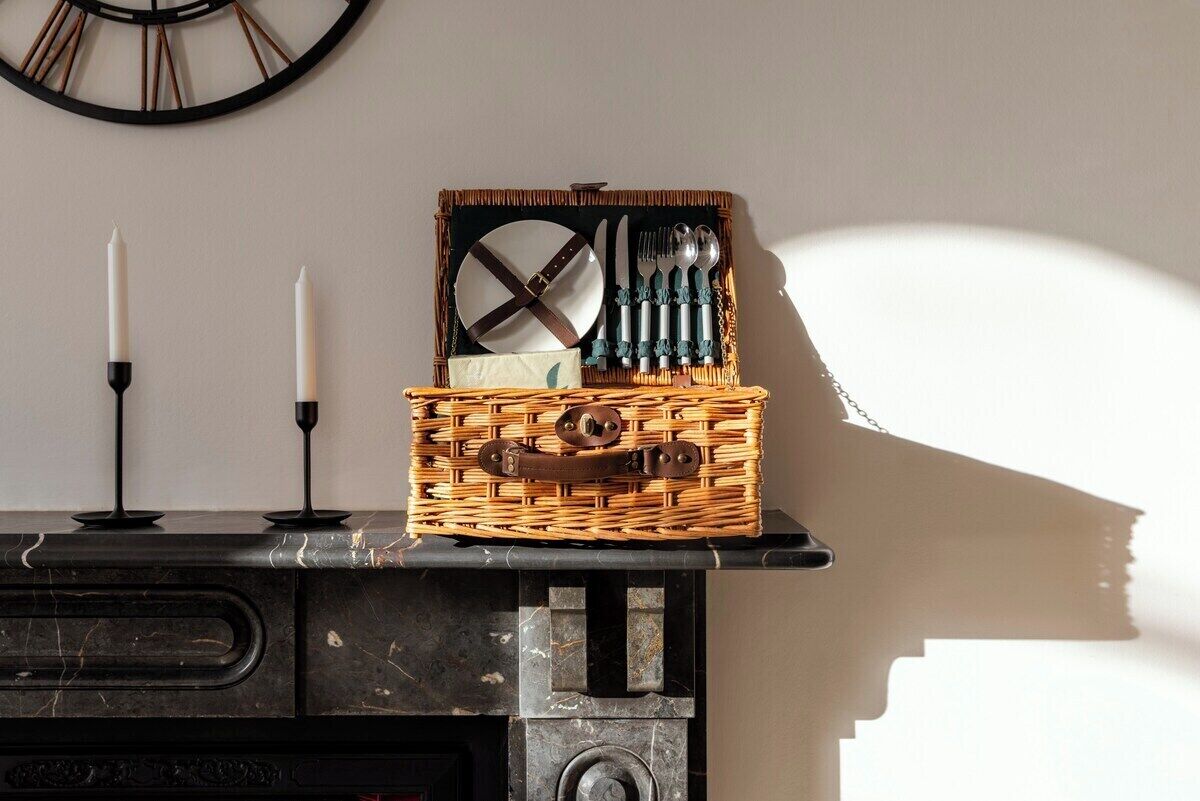 A wicker basket with utensils and a plate on a marble surface, with a clock on the wall behind.