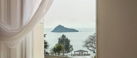 Vista desde una ventana con cortinas blancas, que da a un mar tranquilo y una pequeña isla.
