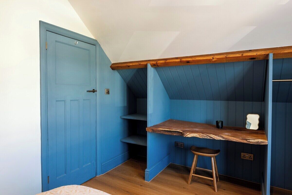 A room with a blue door, a wooden desk, a stool, and a slanted ceiling.