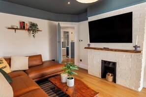Ein Wohnzimmer mit einem braunen Ledersofa, einem Flachbildfernseher über einem weißen Kamin aus Ziegelsteinen und einem Couchtisch mit einer Pflanze.