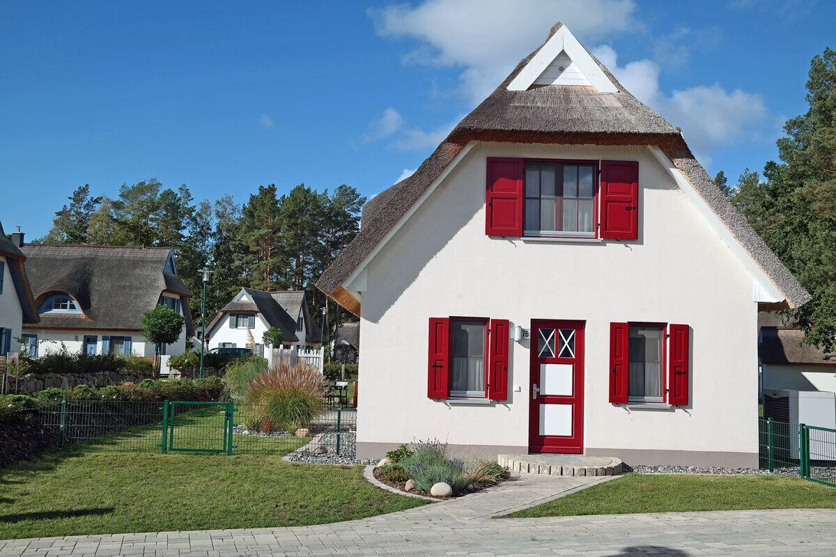 A white house with red shutters, a thatched roof, and a small garden.