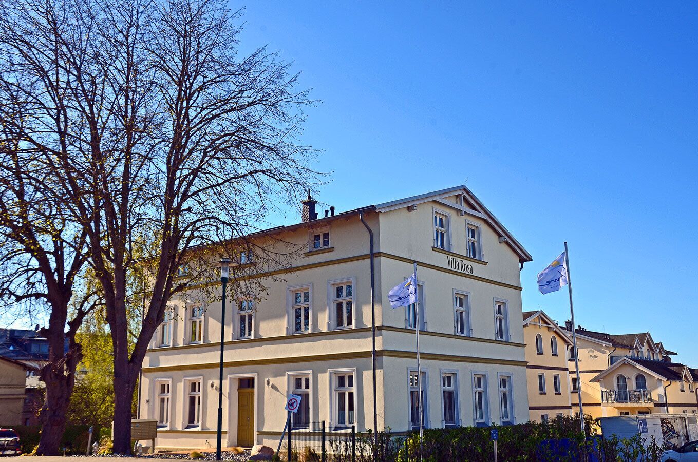 Ein zweigeschossiges Gebäude mit der Aufschrift 'Villa Rosa' an der Fassade, zahlreichen Fenstern und Flaggen auf Fahnenmasten.