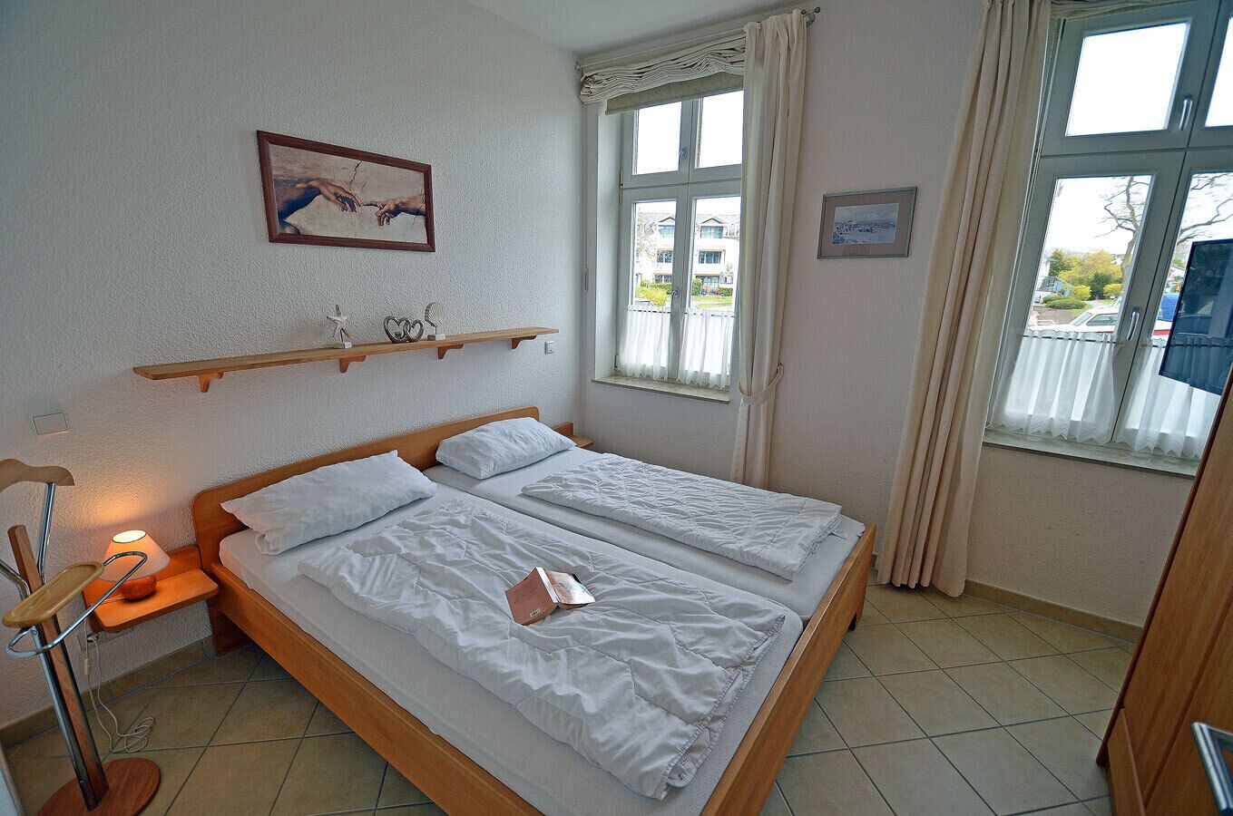 Ein Zimmer mit einem Bett, einer Nachttischlampe, einem gerahmten Bild an der Wand und einem Fenster mit Vorhängen.