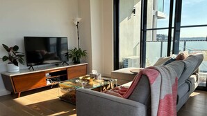 A modern living room with a flat-screen TV, a glass coffee table, a sofa, and a large window offering a view of a body of water and buildings. - New & Quiet 2BR/2BA with Sydney Tower Views, Close to Metro & Shopping Centre (Macquarie Park)