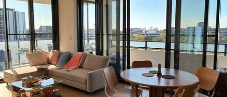 A modern living room with a sofa, coffee table, and dining table. Large windows offer a view of the cityscape.