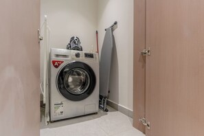 A compact, white front-loading washing machine with a Samsung logo, a grey drying rack, and a wooden door. - HAVN - NEW 1 Bedroom in Neva Residence, JVC (Dubai)
