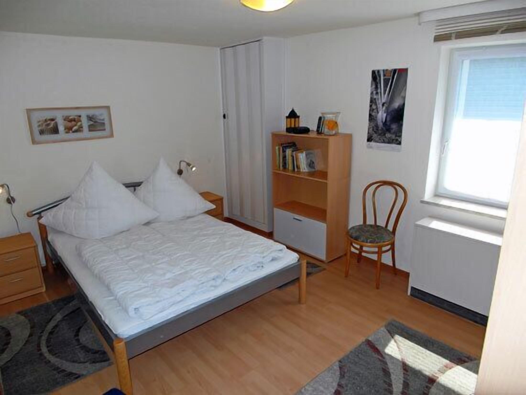 A bedroom with a bed, a chair, a bookshelf, and a window.