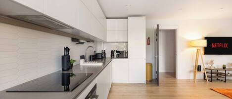 A modern kitchen with white cabinetry, a black countertop, and a built-in oven. There is a microwave, kettle, and coffee maker on the counter. A fire extinguisher is mounted on the wall.