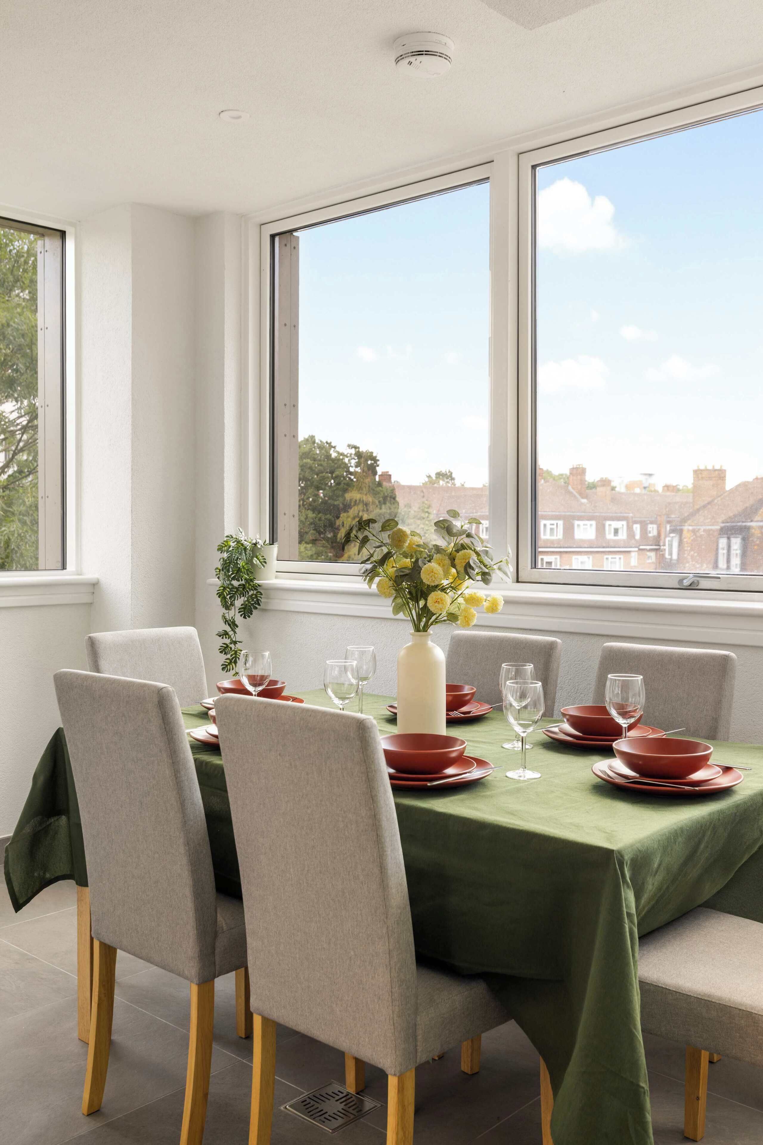 A dining area with a table set for four, chairs, a vase with flowers, and a view of the city through large windows.