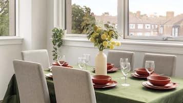 A dining area with a table set for four, chairs, a vase with flowers, and a view of the city through large windows.