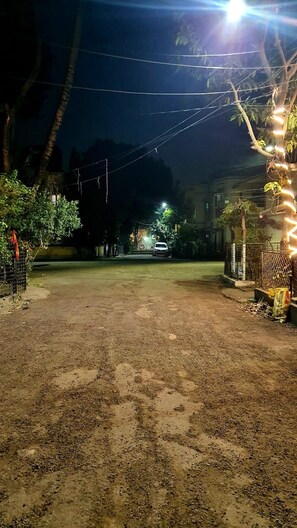 A nighttime view of a residential street with a dirt road, houses, and streetlights.