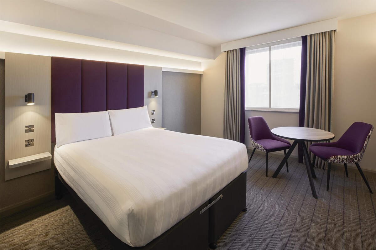 A hotel room with a bed, bedside tables, a desk, two chairs, and a window with curtains.