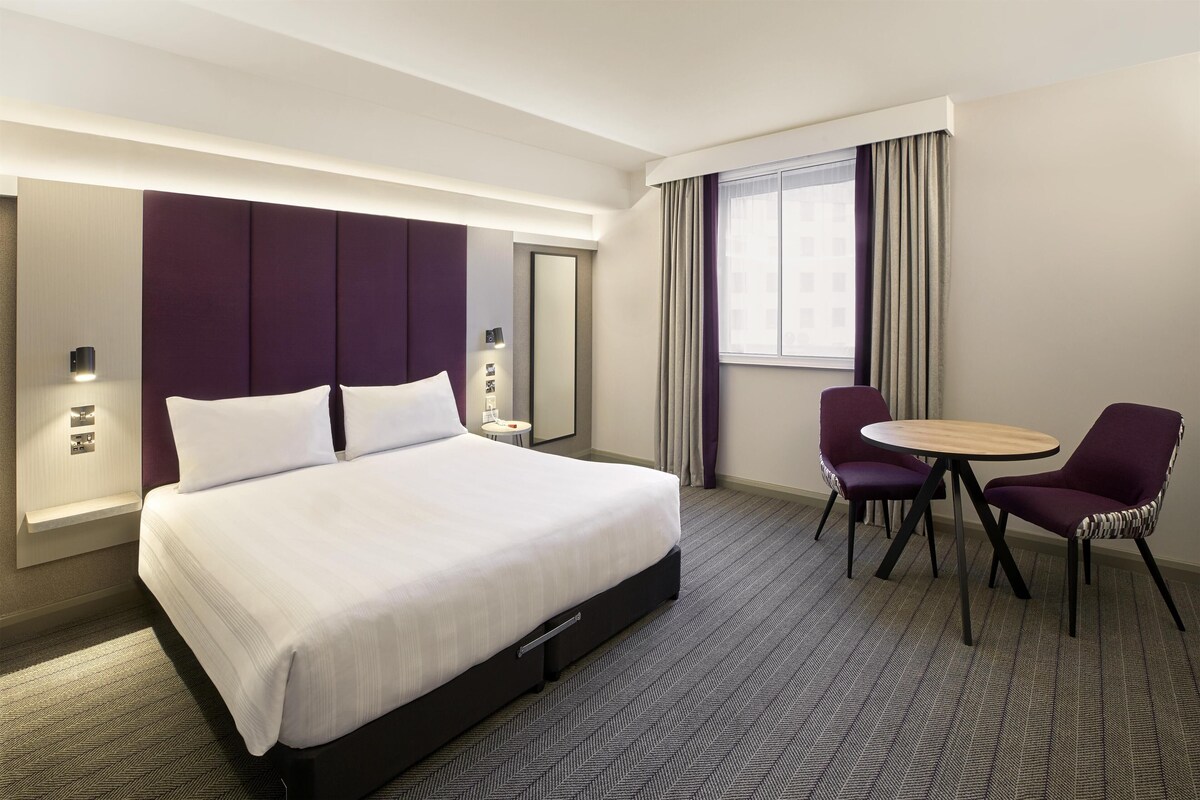 A hotel room with a large bed, a round table, two chairs, a mirror, and a window with curtains.