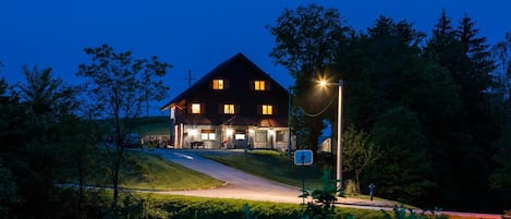 Ein großes, dunkel gefärbtes Haus mit vielen Fenstern und einer gut beleuchteten Veranda, umgeben von Bäumen und einer Rasenfläche.