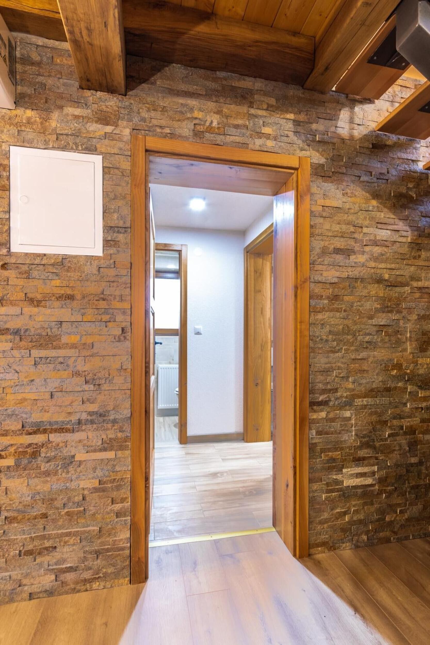 A hallway with a wooden door leading to a bathroom. The walls are made of stone, and there is a wooden ceiling.