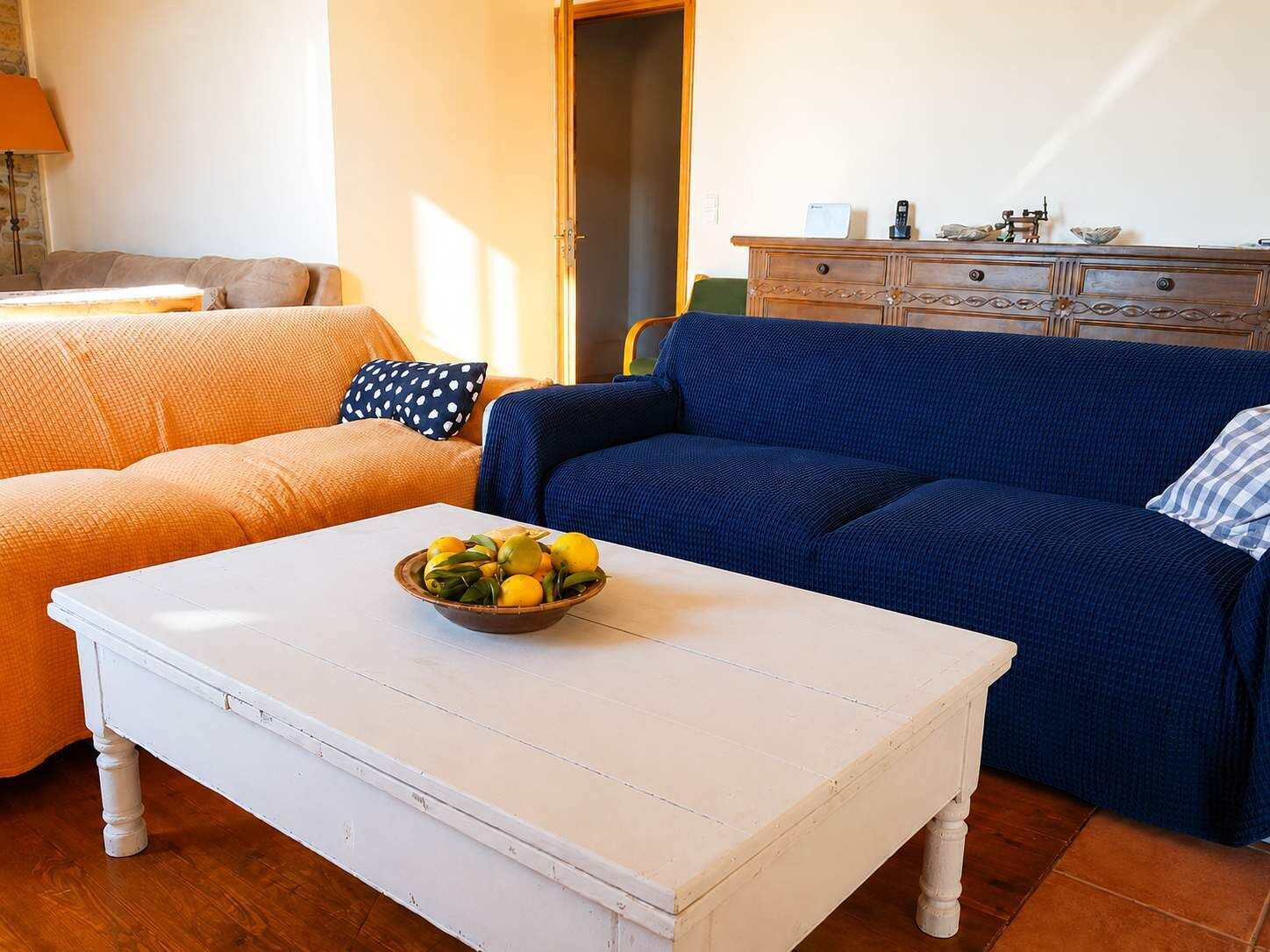 Een woonkamer met twee banken, één oranje en één blauw, een witte salontafel met een schaal fruit, en een houten kast met decoratieve objecten.