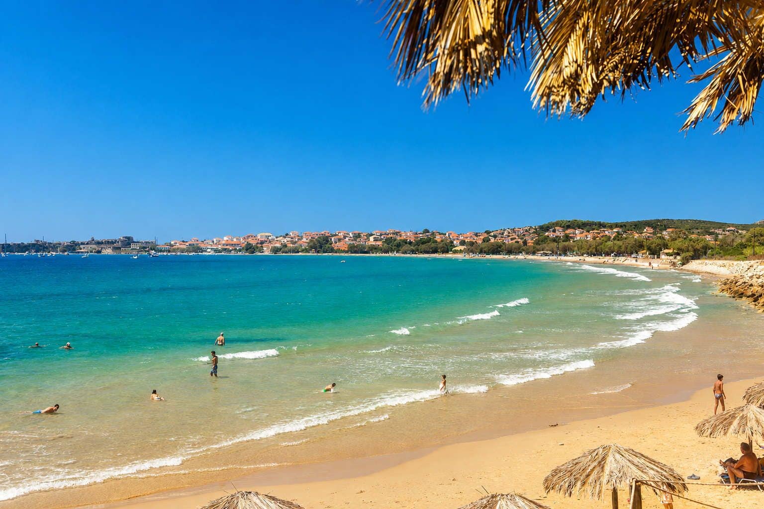 Una playa con aguas turquesas cristalinas, gente nadando y tomando el sol, y un pueblo al fondo.