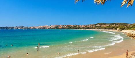 A beach with clear turquoise water, people swimming and sunbathing, and a town in the background.