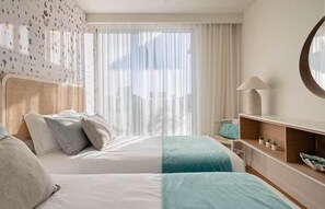 A hotel room with two beds, a bedside table with a lamp, a wall-mounted shelf with plants, and a large window with sheer curtains. - beachfront bliss & breeze (Armação de Pêra)