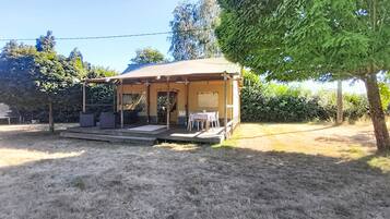 Een overdekt terras met tafels en stoelen, een houten terras en uitzicht op een tuin met bomen en een haag.