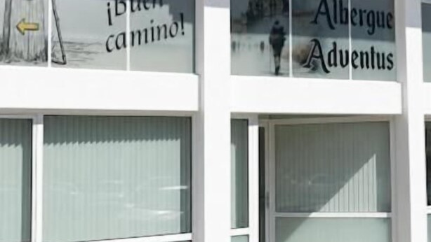 Fachada de edificio con grandes ventanales y señalización que indica '¡Buen Camino!' y 'Albergue Aventus'.