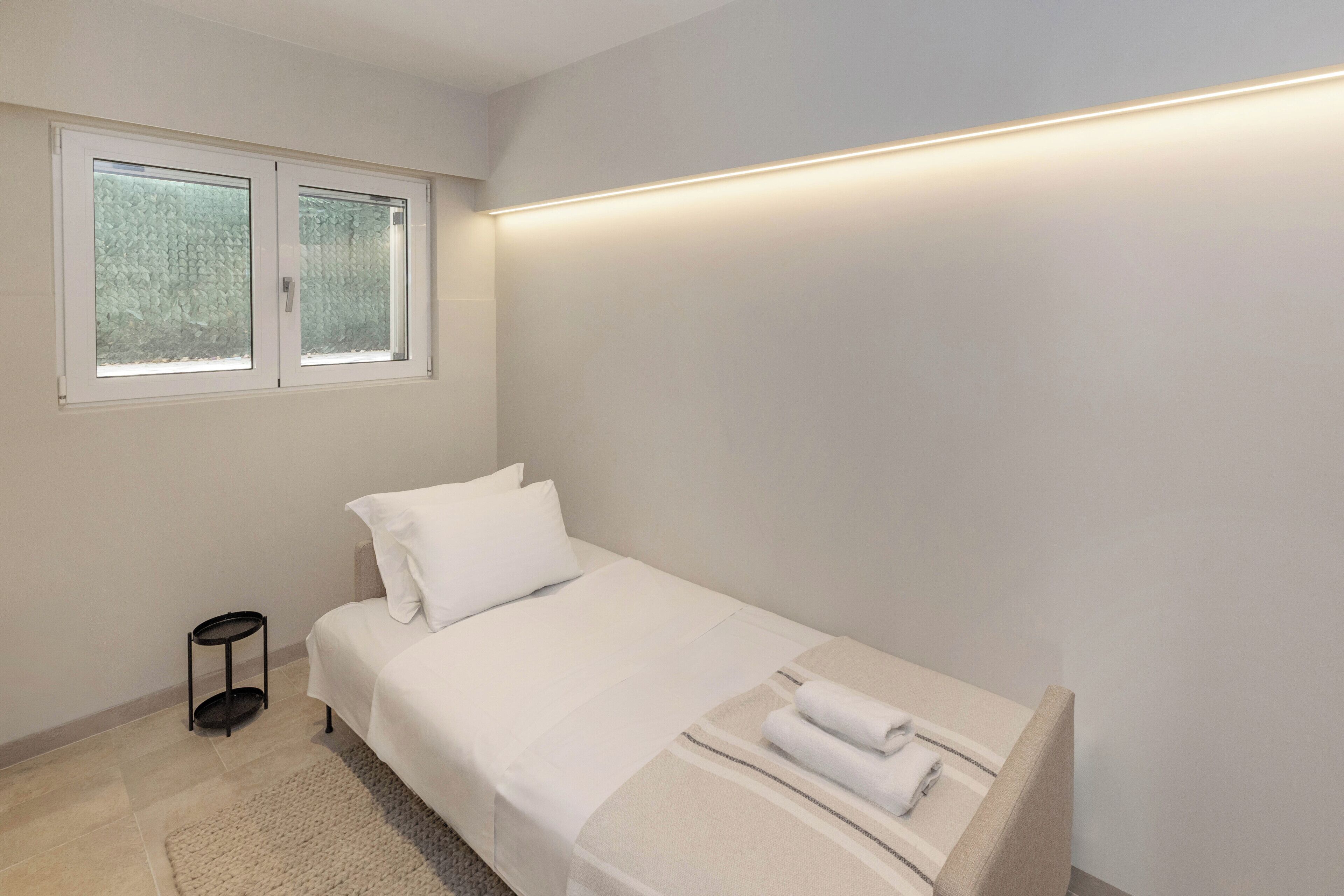 A minimalist bedroom with a single bed, a small bedside table, a window with frosted glass, and a wall-mounted light fixture.