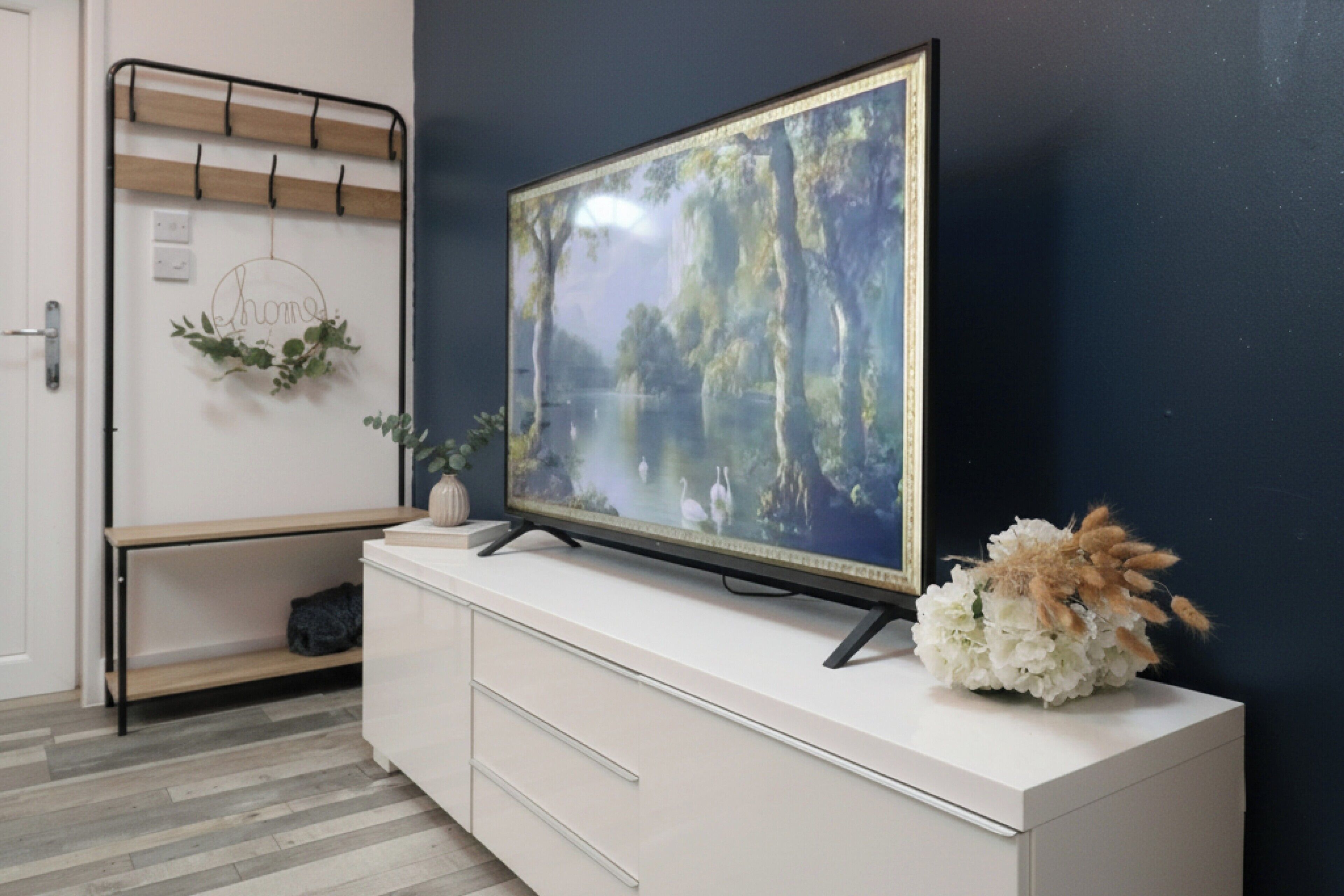 A flat-screen TV on a white cabinet with a forest scene, a coat rack with a hanging garment, and a small plant.
