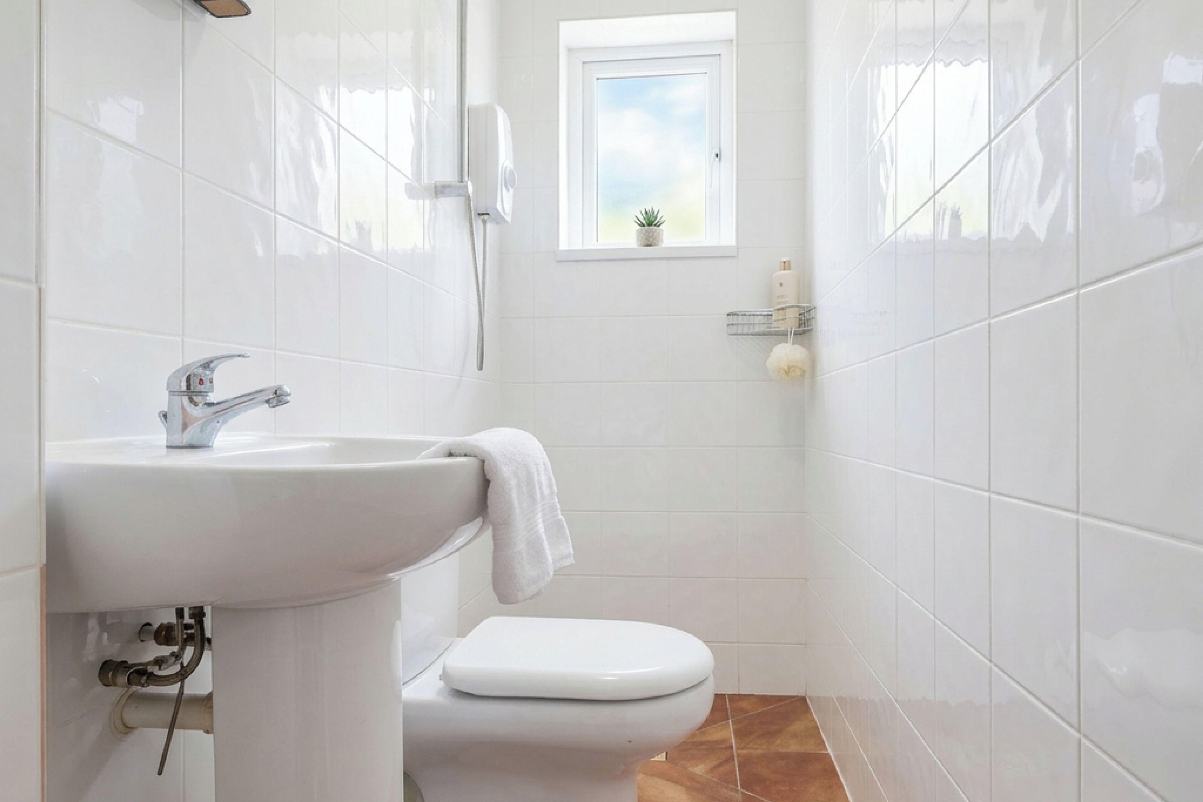 A bathroom with a white sink, toilet, and shower. There is a small window above the toilet.