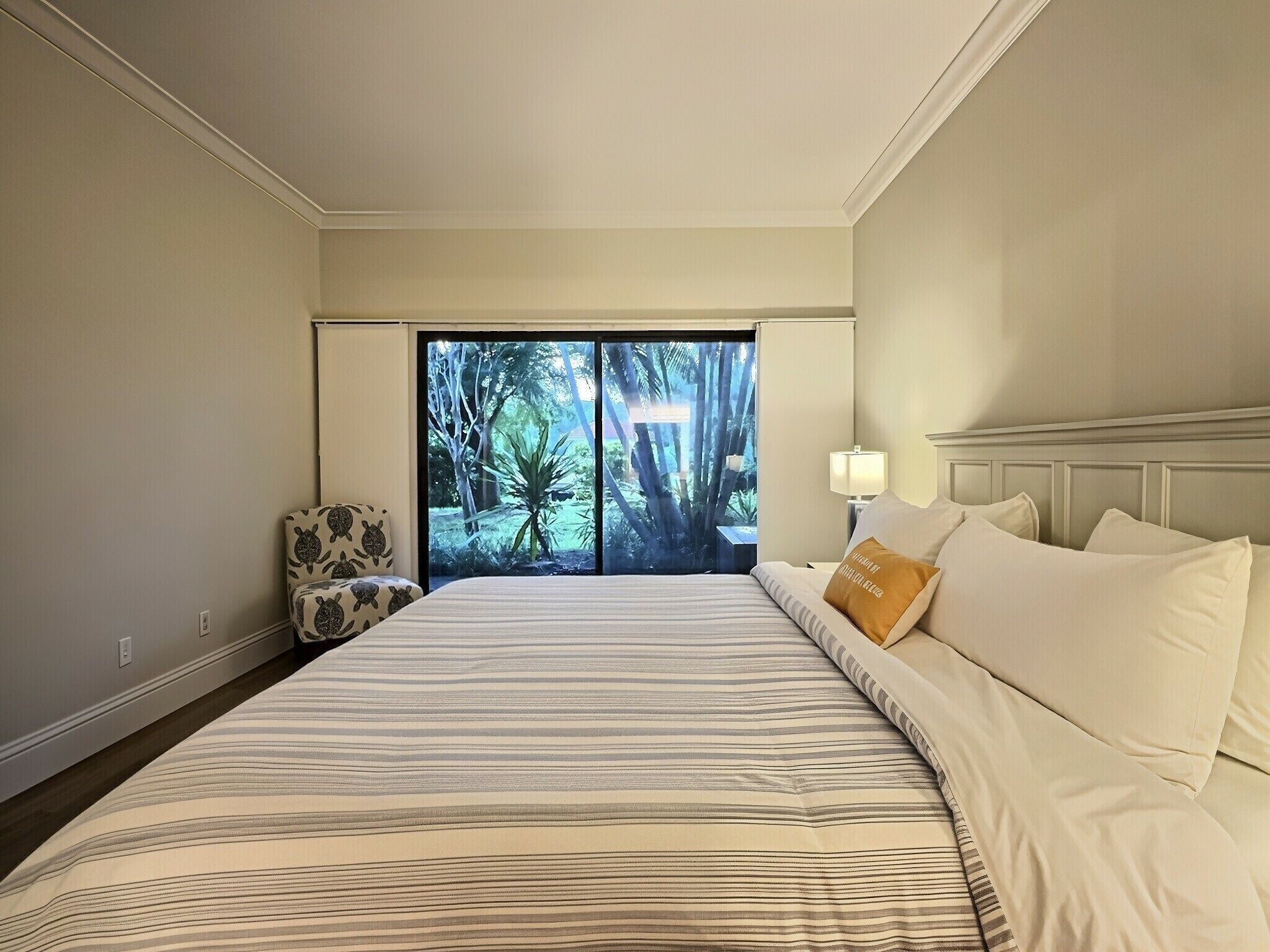 A hotel room with a large bed, a chair, a nightstand, and a view of a tropical landscape through the window.
