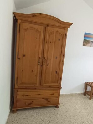Ein Holzkleiderschrank mit zwei Türen und einem unteren Fach, vor einer schlichten Wand, in der Nähe steht ein kleiner Hocker.