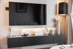 A modern living room with a flat-screen TV mounted on the wall, a dark-colored console table with decorative items, and a floor lamp. - StayShort - Meydan One sleeps 4 close to Meydan Racecourse! (Sobha Hartland)