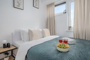 A hotel room with a bed, pillows, a bedside table with a lamp, a window with curtains, and a bowl of fruit on the bed. - Lumina 2-room Apart near Warsaw Center (Warszawa)