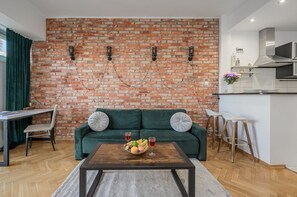 A modern living room with a brick wall, a green sofa, a wooden coffee table with fruit, and a kitchen area with white cabinets and a stainless steel range hood. - Lumina 2-room Apart near Warsaw Center (Warszawa)