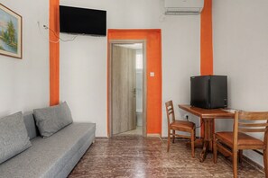 A room with a grey sofa, a wooden table with a black television, and an orange accent wall. - Spacious Room Next to Kalamata Airport, Heart of Messinia (Mikromani)