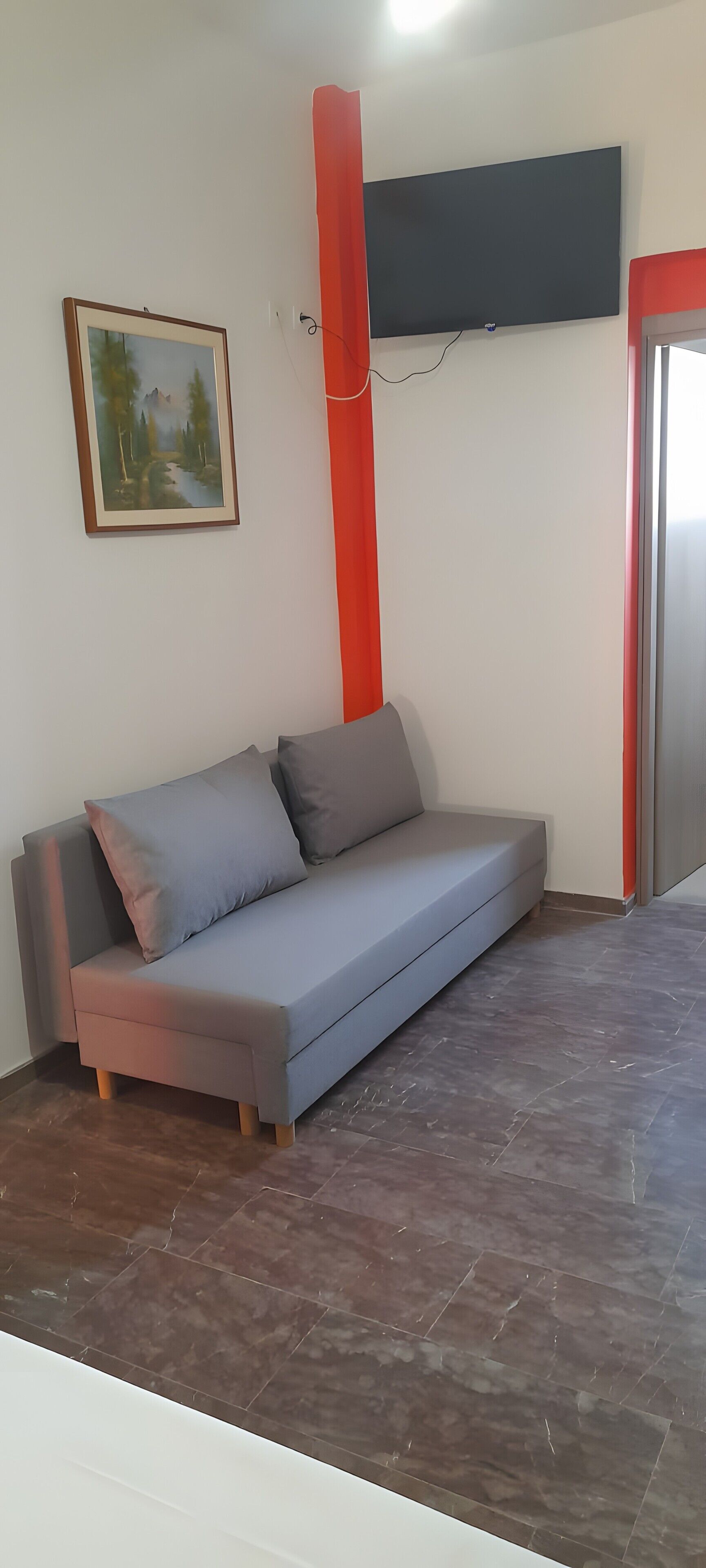 Een kamer met een grijze bank, een schilderij, een rode zuil en een flatscreen televisie aan de muur.