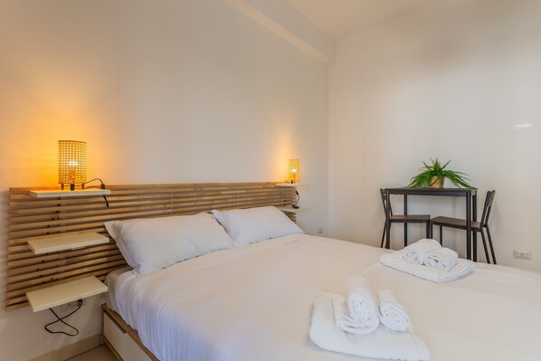 Een moderne hotelkamer met een houten hoofdeinde, een bed met witte lakens, twee bedlampen, een kleine tafel met een plant en een stoel.