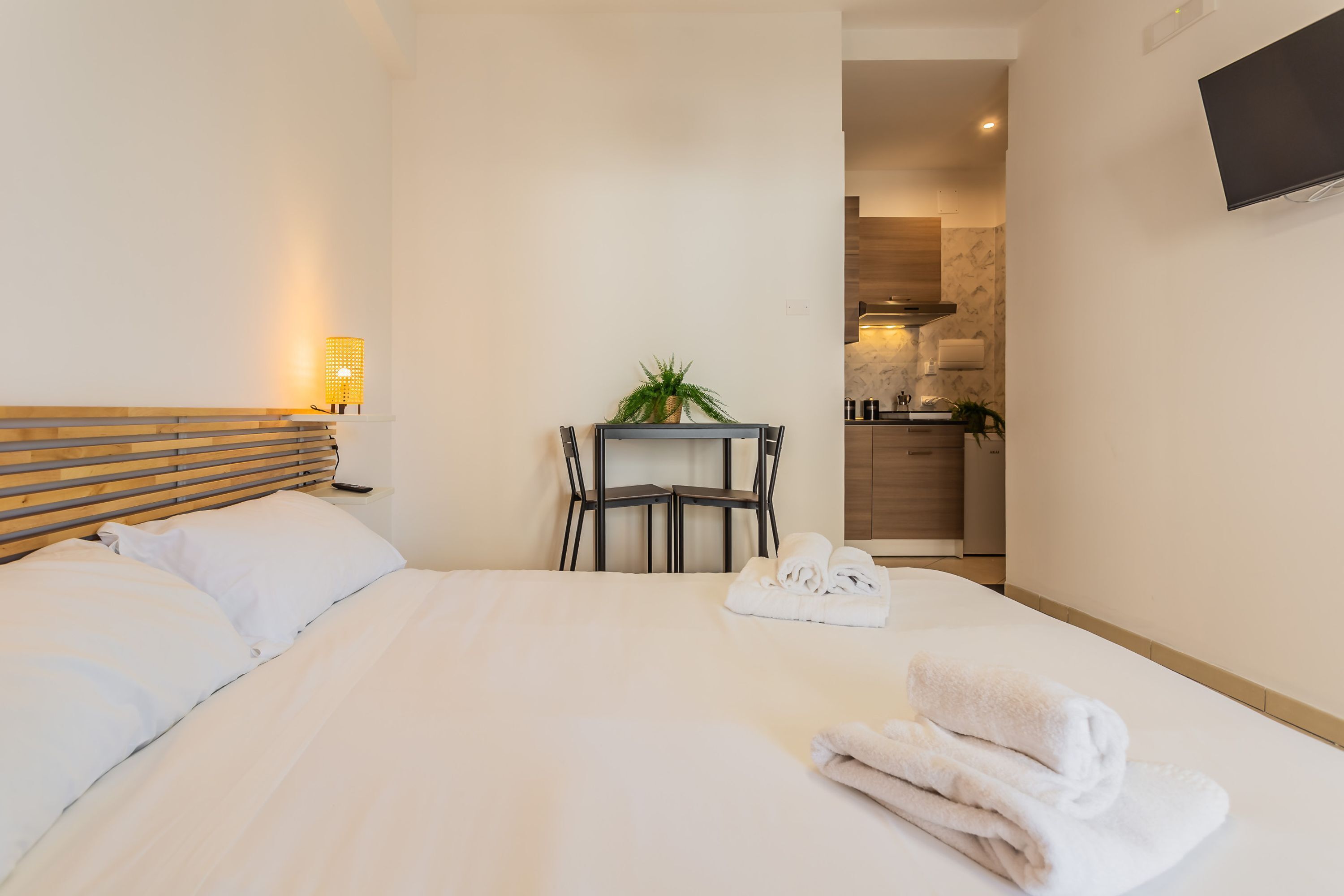 Quarto de hotel com uma cama, mesa de cabeceira, abajur e vista para a cozinha.