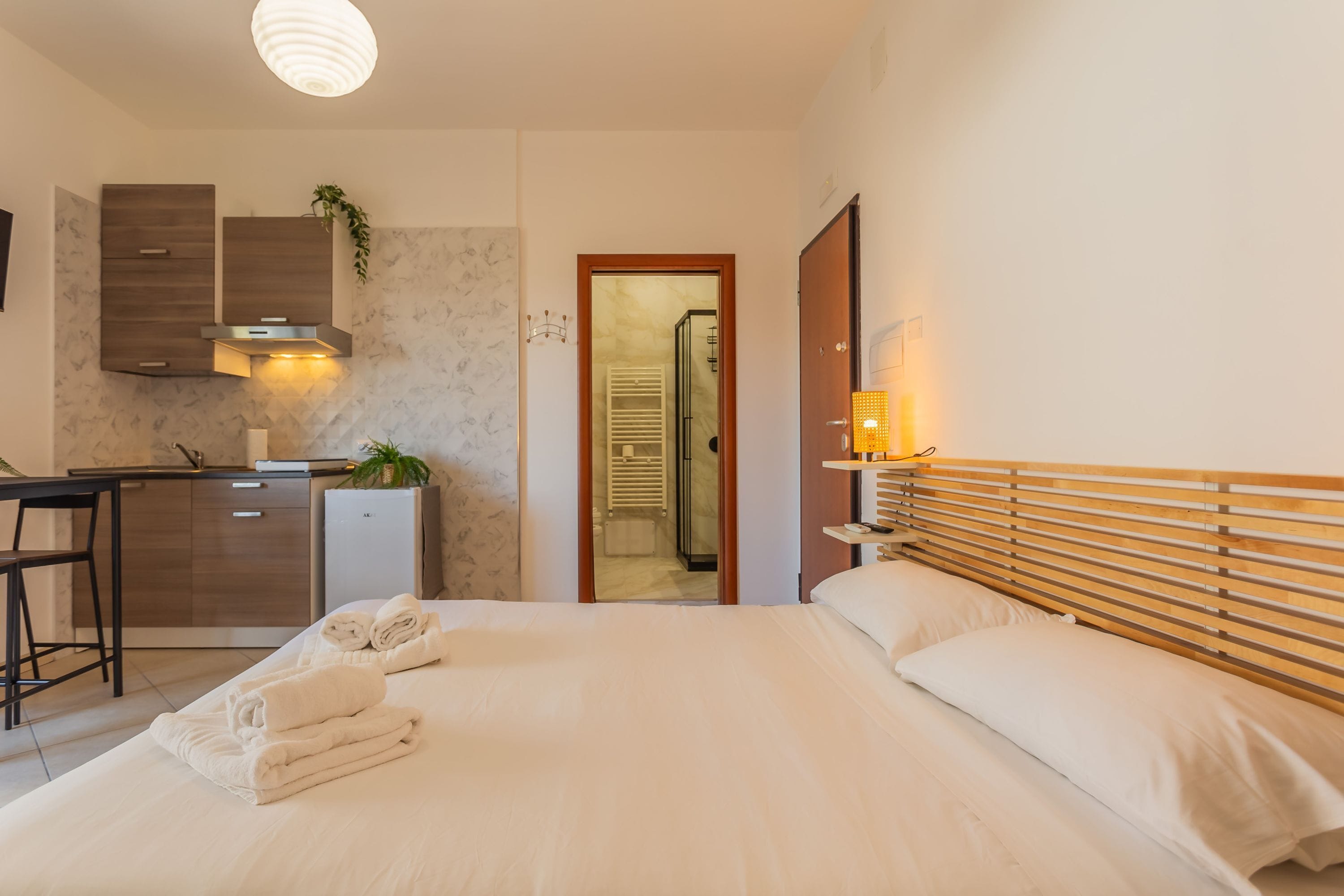 A hotel room with a bed, a desk, a kitchenette, and a bathroom visible through an open door.