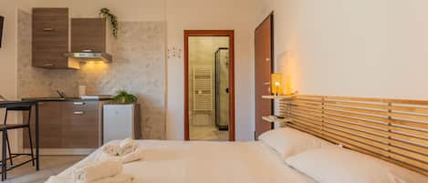 A hotel room with a bed, a desk, a kitchenette, and a bathroom visible through an open door.