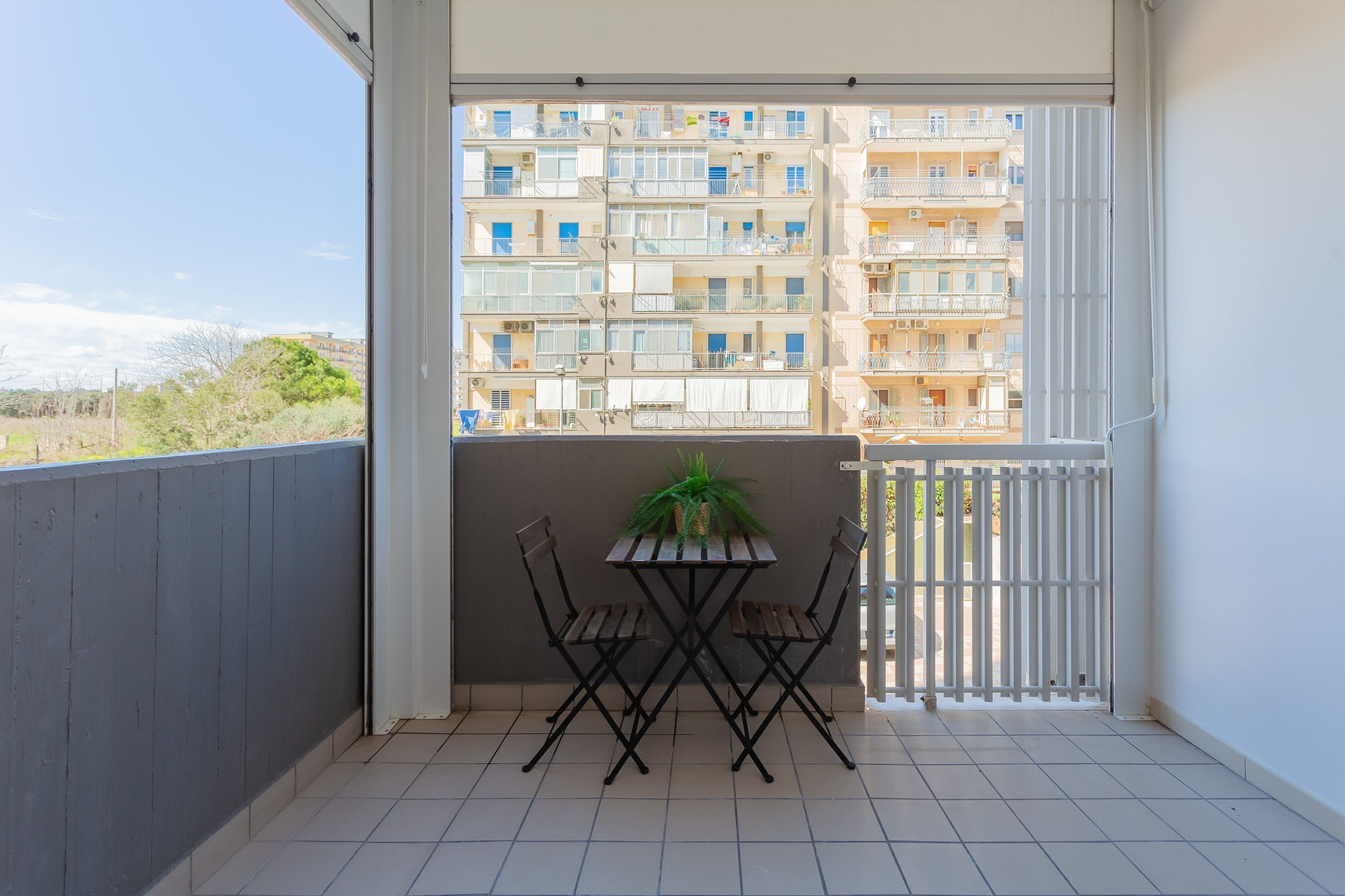 Un balcon avec une table et des chaises, un pot de fleurs et une vue sur un immeuble à plusieurs étages.