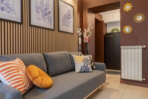 Ein modernes Wohnzimmer mit grauem Sofa, dekorativen Kissen, gerahmten Bildern an der Wand und einer Tür mit Spiegel und Sonnendekoration.