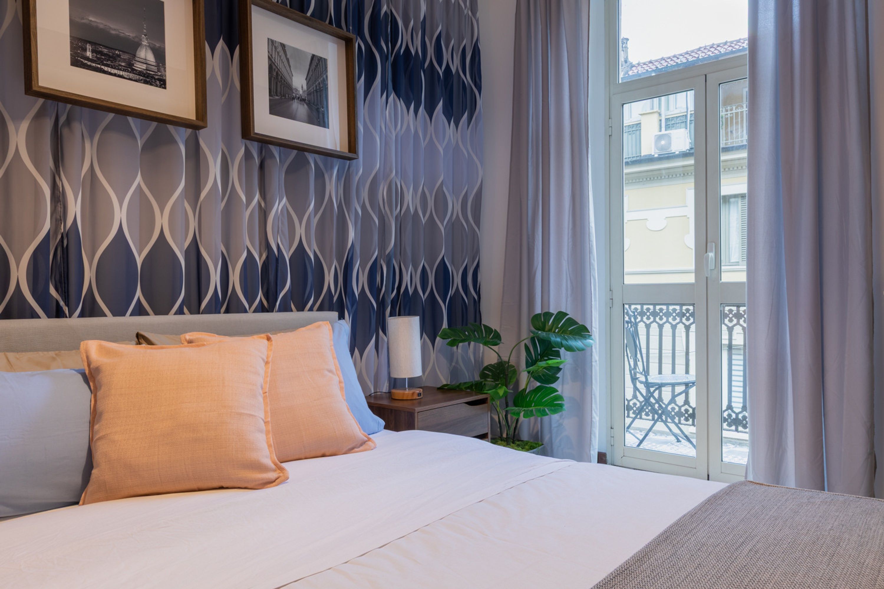 A hotel room with a bed, two framed pictures on the wall, a nightstand with a lamp, and a view of the outside balcony.